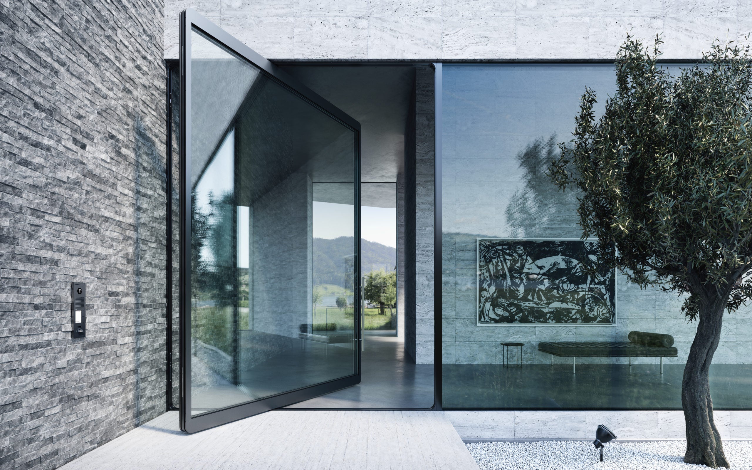 Modern architectural design featuring a large, angular pivot door made of glass, set within a minimalist home facade. The door is open, leading into a polished interior space that includes a wall-mounted artwork. The exterior showcases a textured stone wall and a neatly landscaped area with a mature olive tree and small pebbles, under a clear sky with distant hills visible, emphasizing the blend of indoor and outdoor living.